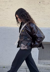 Veste en cuir marron brillant portée sur un sweat-shirt polaire gris à motif camouflage, pantalon noir, avec un sac à bandoulière clair, marchant sur un trottoir.