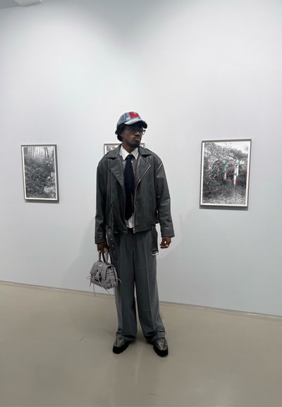 Chaqueta de biker de cuero gris sobre una camisa blanca y corbata oscura, combinada con pantalones grises de pierna ancha. Lleva un pequeño bolso gris. Gorra de mezclilla con detalles rojos.