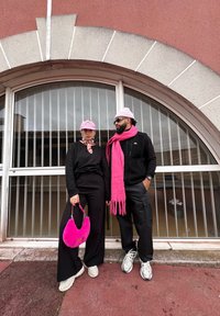 Δύο άτομα στέκονται δίπλα σε ένα παράθυρο. Ο ένας φοράει μαύρα ρούχα και κρατάει μια ροζ τσάντα από γούνα. Ο άλλος φοράει μια μαύρη γιλέκο και ροζ κασκόλ.