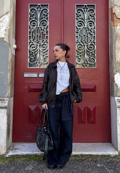 Mujer con chaqueta negra y blusa blanca estampada, sosteniendo una bolsa negra, de pie frente a una puerta roja con rejas de metal decorativas.