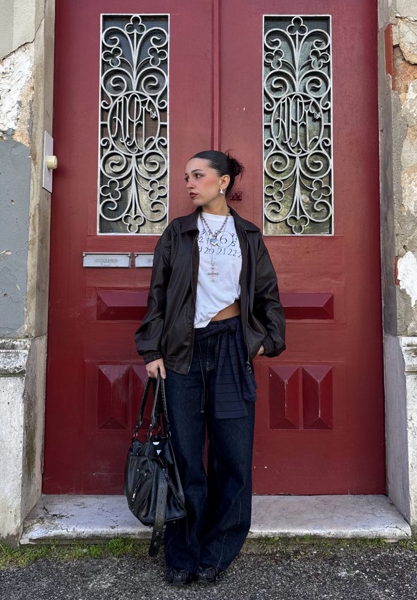 Γυναίκα που φοράει μαύρο μπουφάν και λευκό μπλουζάκι με στάμπα, κρατώντας μαύρη τσάντα, στέκεται μπροστά σε κόκκινη πόρτα με περίτεχνα μεταλλικά κάγκελα.