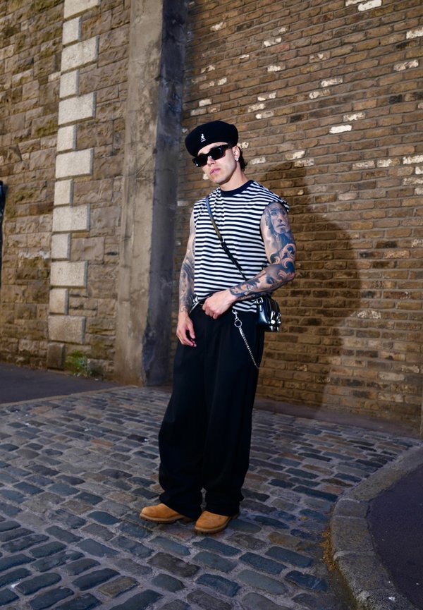 Mężczyzna z tatuażami, noszący czarny beret, pasiastą koszulkę bez rękawów, czarne spodnie, okulary przeciwsłoneczne i jasnobrązowe buty stojący na brukowanej ulicy obok ceglanej ściany.