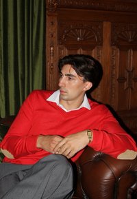 Roter Pullover mit weißem Kragen, graue Hose, sitzt auf einem braunen Lederstuhl, vor grüner Gardine und kunstvollem Holzhintergrund.