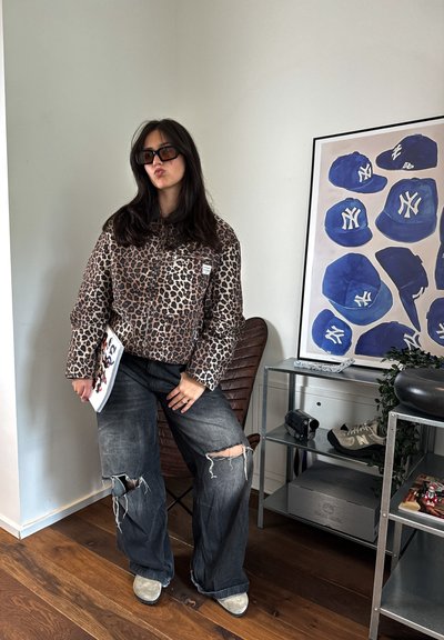 Chaqueta acolchada de estampado de leopardo, jeans grises oversize desgastados y botas grises. Los accesorios incluyen gafas de sol negras y una revista.