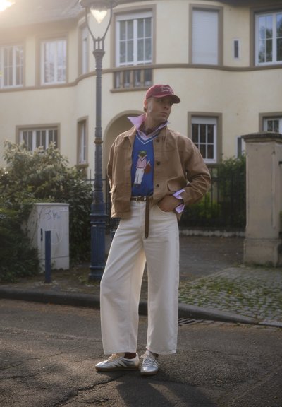 Persona con chaqueta color beige, suéter azul con gráfico, pantalones blancos de pierna ancha, gorra roja y zapatillas plateadas, de pie en una calle cerca de un poste de luz.