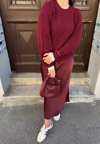 Un pull texturé bordeaux, une longue jupe bordeaux, des baskets blanches avec des rayures marron foncé et un sac à main tissé marron foncé sont portés par une personne.