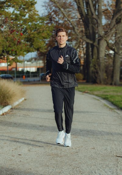 Chaqueta ligera de running negra con acentos reflectantes, pantalones de running negros y zapatillas deportivas blancas. Camino de grava con vegetación de fondo.