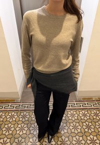 Beige stickad tröja kombinerad med en mörkgrå omlott och svarta vida byxor. Mjuk textur och enkel design utan synliga mönster.