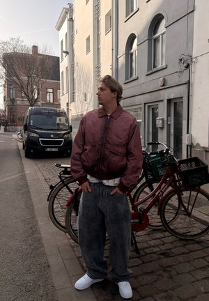 Joven con chaqueta granate y vaqueros holgados de pie en una calle adoquinada cerca de una bicicleta roja estacionada y una furgoneta negra junto a edificios residenciales.