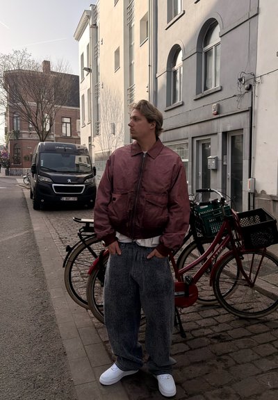 Joven con chaqueta granate y vaqueros holgados de pie en una calle adoquinada cerca de una bicicleta roja estacionada y una furgoneta negra junto a edificios residenciales.