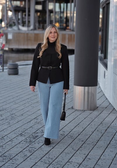 Mujer rubia con chaqueta negra, vaqueros anchos azul claro y botas negras caminando sobre un pavimento con patrón mientras sostiene un pequeño bolso negro.