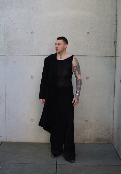 Conjunto de ropa negra que presenta una blusa transparente y una falda larga, complementado por un abrigo largo que atraviesa un hombro y un brazalete plateado.