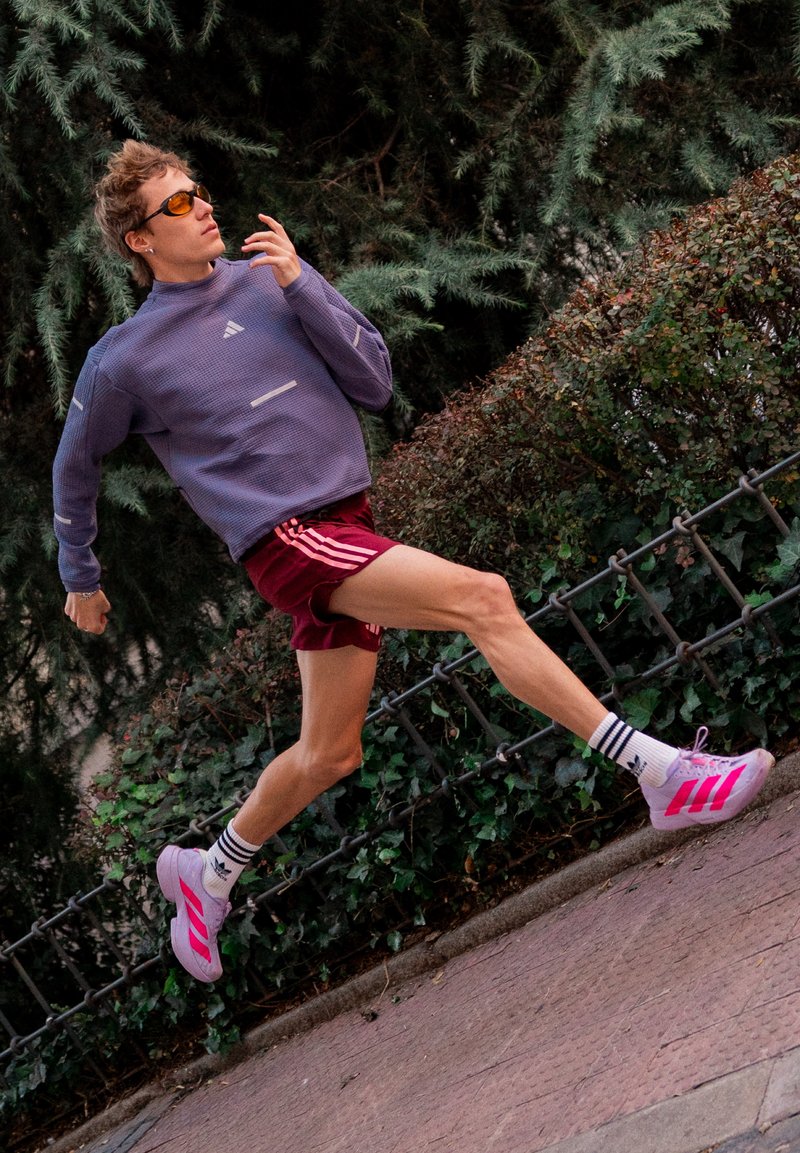 Corridore con felpa viola, pantaloncini color marrone scuro e scarpe rosa salta su un marciapiede accanto a una recinzione metallica con cespugli verdi e alberi.