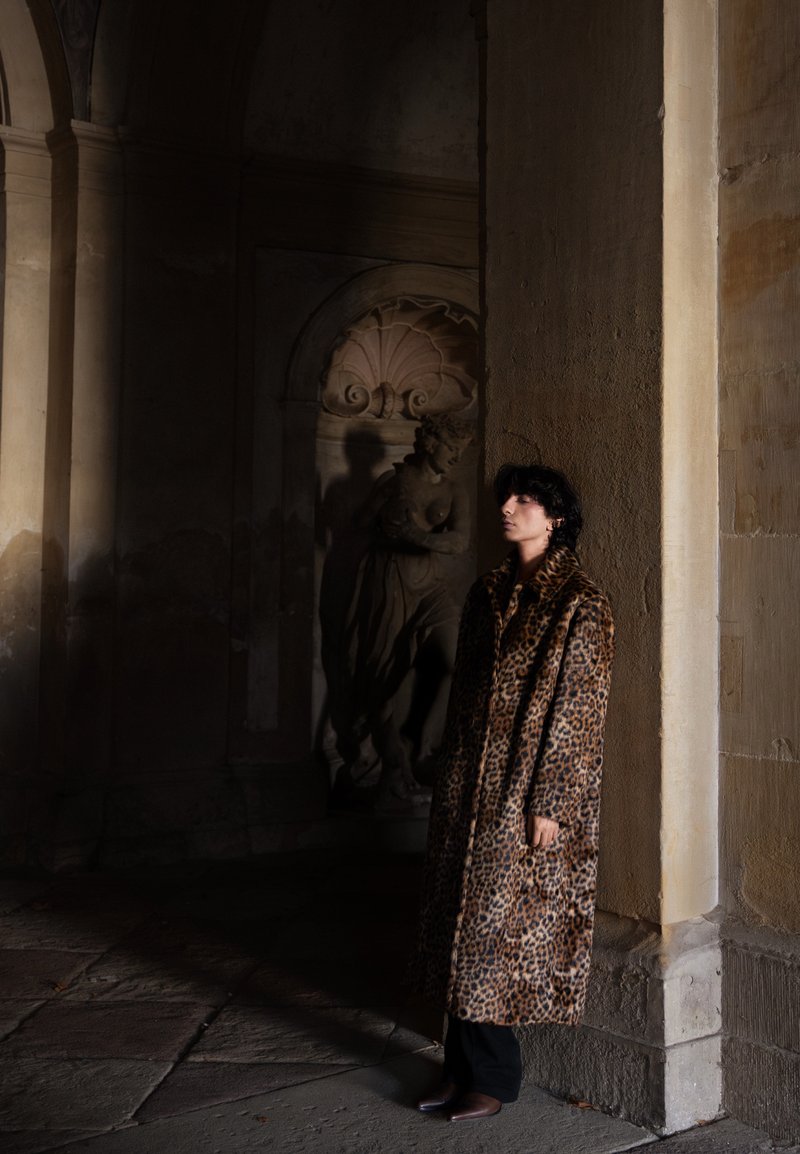 Personne en manteau à imprimé léopard appuyée contre un pilier en pierre dans un couloir historique faiblement éclairé avec une statue classique en arrière-plan.