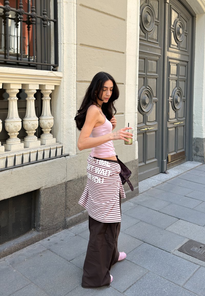 Jeune femme en débardeur rose et jupe rayée tenant un verre, debout sur un trottoir urbain près de portes grises ornées et d'une balustrade en pierre.