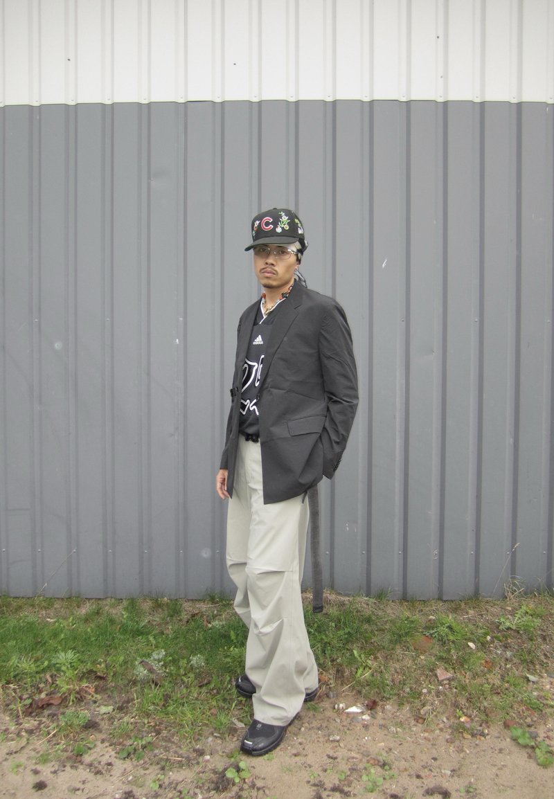 Black blazer over a black shirt with a white graphic, cream-colored wide-leg pants, black shoes, and a black cap with green accents.