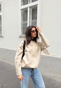 Svetlo bež oversized jakna z ostrim ovratnikom, v kombinaciji s svetlo modrimi jeans hlačami. Črne sončna očala in črna torbica s trakovi dopolnjujeta videz.