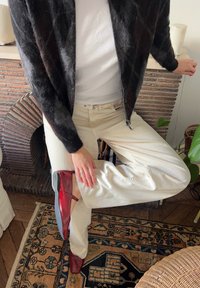 Črn pleteni kardigan, bela majica s kratkimi rokavi, bež hlače in rdeči lakasti čevlji. Roka počiva na kolenu, kar poudarja detajl čevlja.