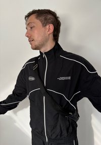 Schwarze, mit Reißverschluss versehene Jacke mit weißen Akzenten und Logo, kombiniert mit einer schwarzen Umhängetasche. Glatte Textur; sportliches Design mit hohem Kragen.