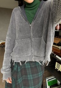 Personne portant un col roulé vert, un pull tricoté gris à fermeture éclair avec des bords effilochés, et une jupe à carreaux vert foncé et noir, à l'intérieur près des étagères.