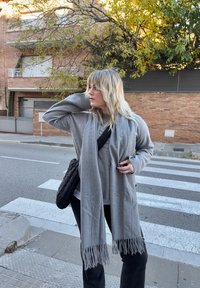 Femme blonde en pull gris et écharpe se tient près d'un passage piéton, regardant sur le côté avec une main derrière la tête et l'autre tenant l'écharpe.