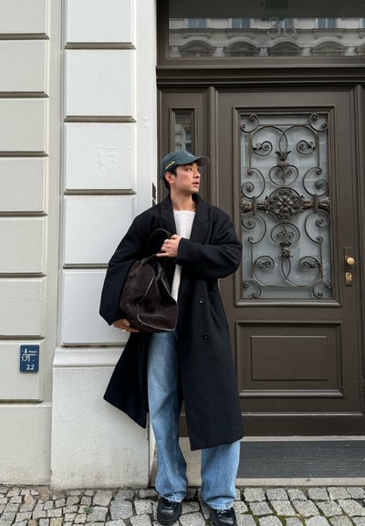 Abrigo negro largo sobre un suéter crema, jeans azul claro y zapatos negros. Sostiene un bolso marrón oscuro y lleva una gorra verde. El fondo presenta una puerta decorativa.