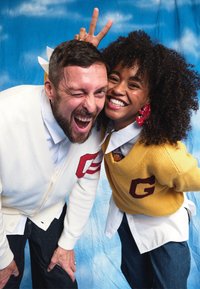 Two people wearing contrasting cardigans: one white with red lettering and stripes, the other yellow with burgundy lettering. They are wearing denim bottoms and smiling.