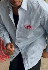 Camicia a righe blu e bianca con un grande logo ricamato rosso sul petto a sinistra. Tessuto in cotone, vestibilità rilassata.