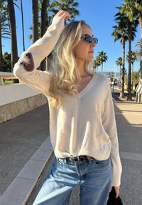Pull beige en tricot avec un col en V et des manches longues, orné d'un écusson marron sur le bras gauche. Associé à un jean bleu clair.