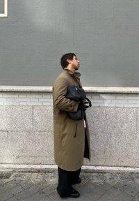 Μακρύ μπεζ παλτό, μαύρη τσάντα ώμου κρατημένη, σκούρα παντελόνια και παπούτσια. Στεκόμενος μπροστά σε ένα γκρι τοίχο, προφίλ άποψη. Διακριτές υφές και τόνους.