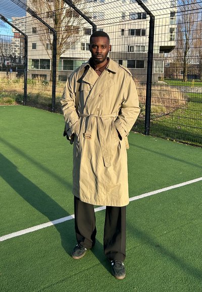 Trench beige con cintura ceñida, grandes solapas y bolsillos, combinado con pantalones de pierna ancha negros y zapatos oscuros, en un campo de césped verde.