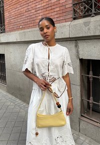 Blusa bianca ricamata con maniche corte, abbinata a una gonna bianca floreale. Tiene una borsa di pelle gialla decorata con perline.