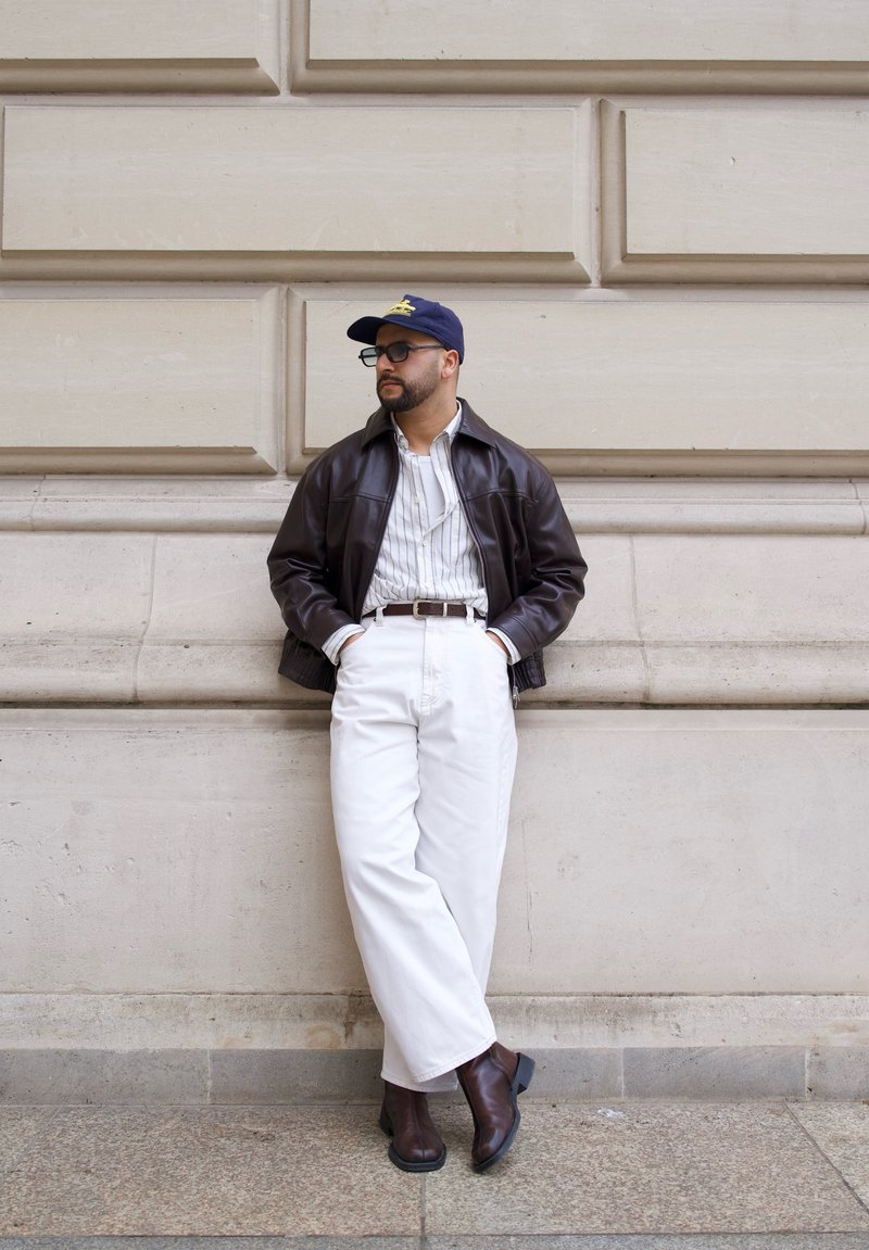 Άνδρας ακουμπάει σε φωτεινό λιθόκτιστο τοίχο, φοράει σκούρο δερμάτινο μπουφάν, ριγέ πουκάμισο, λευκό παντελόνι, καφέ μπότες, μπλε καπέλο και γυαλιά.