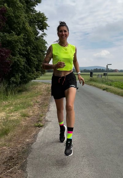 Top corto de tirantes amarillo neón, pantalones cortos negros, calcetines multicolores (rosa, verde, amarillo) con zapatillas negras, corriendo por una carretera rural.