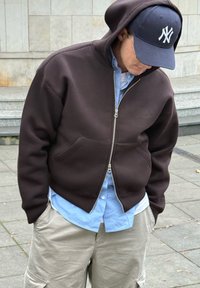 Person wearing a dark brown zip-up hoodie, light blue shirt, beige cargo pants, and a navy blue cap with a white logo, standing outdoors.
