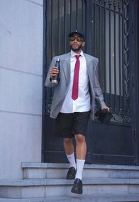 Ένας άντρας φοράει ένα γκρι oversize σακάκι, λευκό πουκάμισο, κόκκινη γραβάτα, μαύρες βερμούδες, μαύρες παπούτσια και λευκές κάλτσες. Κρατάει ένα μπουκάλι νερού από ανοξείδωτο ατσάλι.