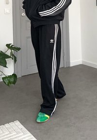 Pantalon de survêtement adidas noir avec des bandes latérales blanches, associé à des chaussures vertes colorées avec un accent jaune et un design à lacets.