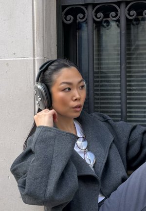 Joven con auriculares grandes y un abrigo gris, ajustando los auriculares mientras está sentado junto a un edificio con una ventana con rejas.