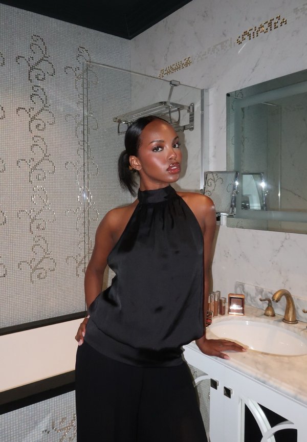 Frau in einem schwarzen ärmellosen Oberteil posiert mit einer Hand auf der Badezimmerablage neben Waschbecken, Spiegel und gefliestem Duschwand.