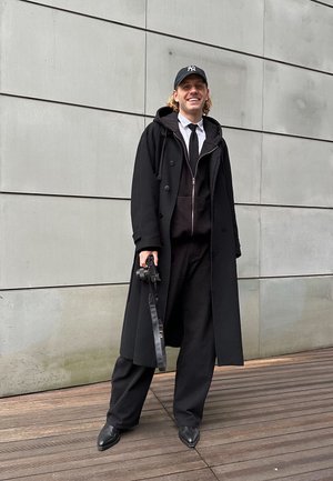 Abrigo de lana negro con capucha, superpuesto a una sudadera negra con cremallera y una camisa blanca. Pantalones negros y cámara en mano. Zapatos negros planos.