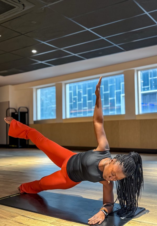 Hosszú fonott hajjal rendelkező nő fekete felsőben és piros nadrágban oldalirányú plank pózban egyensúlyoz egy szőnyegen egy világos fitnesz stúdióban.