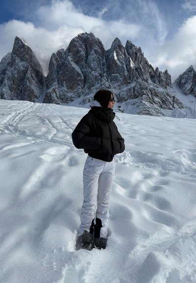 Persona con chaqueta negra y pantalones blancos de pie sobre la nieve con picos montañosos rocosos al fondo bajo un cielo nublado.
