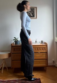 Femme aux cheveux bouclés attachés en chignon, portant un haut bleu clair à manches longues, un jean noir à jambes larges et des bottes à talons noires, debout de profil à l'intérieur.