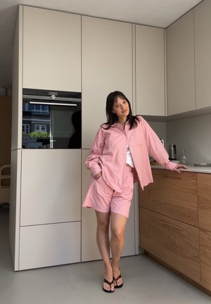 Svetloružová voľná košeľa a zodpovedajúce šortky vyrobené z textúrovanej bavlnenej tkaniny. Jednoduché gombíky, voľný strih a nosené s hnedými sandálmi.