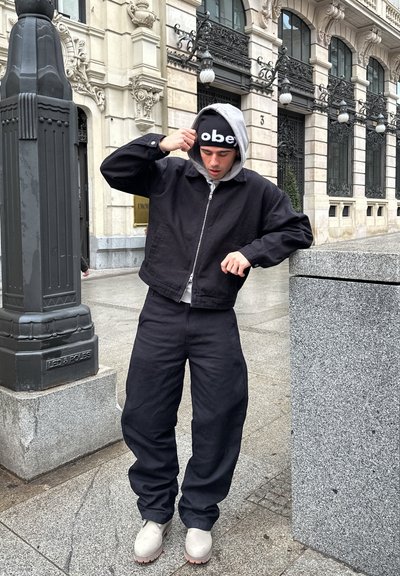 Chaqueta negra corta con capucha gris, combinada con pantalones negros sueltos y zapatos beige. Lleva un gorro negro con el texto "obey".