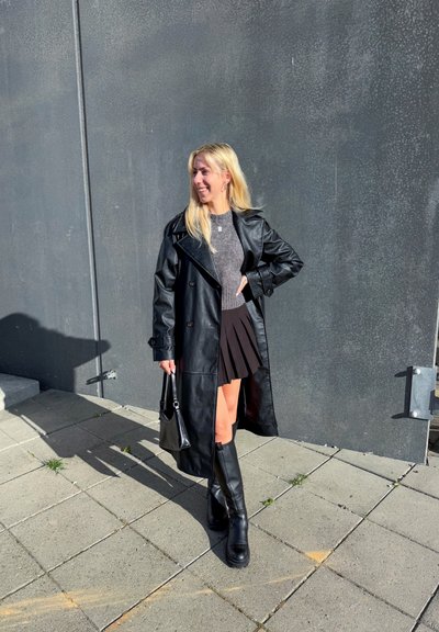 Abrigo de trench de cuero negro sobre un top de punto gris y una falda negra con pliegues. Botas negras hasta la rodilla y un pequeño bolso negro completan el look.