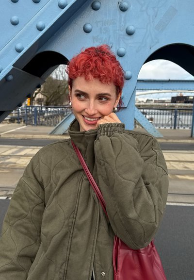 Joven con cabello rizado y rojo sonríe mientras lleva una chaqueta acolchada verde y una bolsa roja, de pie junto a una estructura de puente metálico azul.