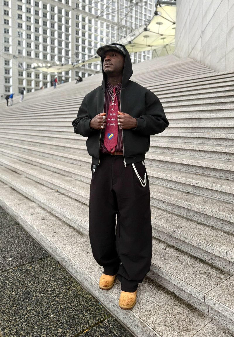 Hombre con sudadera negra con capucha, corbata roja, pantalones anchos negros y zapatos color beige de pie sobre grandes escalones de piedra en un entorno urbano con una fachada de edificio geométrica.
