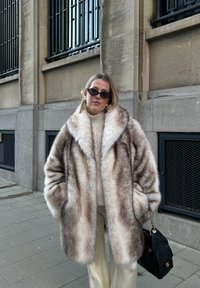 Femme portant un grand manteau en fourrure brun clair, un pull crème, un pantalon beige, des lunettes de soleil noires, et tenant un sac à main noir, debout sur un trottoir en ville.