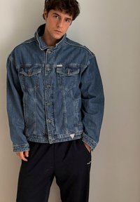 Giacca di jeans blu con collo alto, quattro tasche frontali, chiusura con bottoni sul davanti e un piccolo logo triangolare all'orlo.